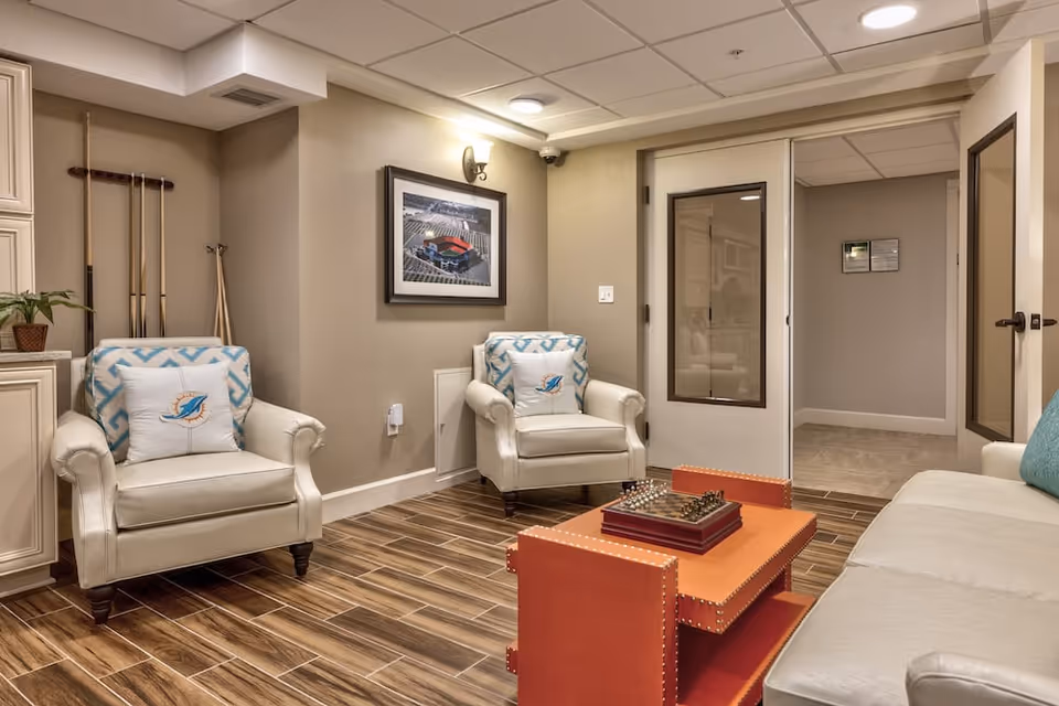 Cozy seating lounge with two upholstered armchairs, a sofa, an orange coffee table, and framed artwork in a well-lit interior room.