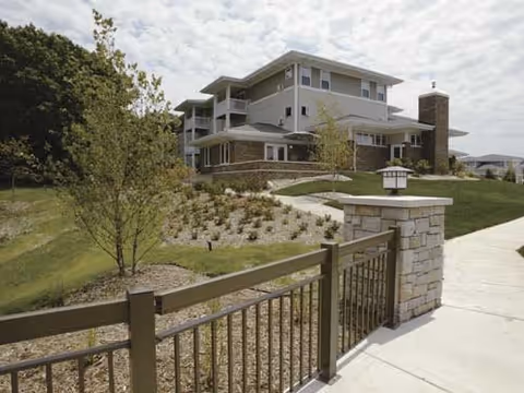 Multi-story senior living building with landscaped grounds, a paved walkway, and a stone-and-wood fence in the foreground.