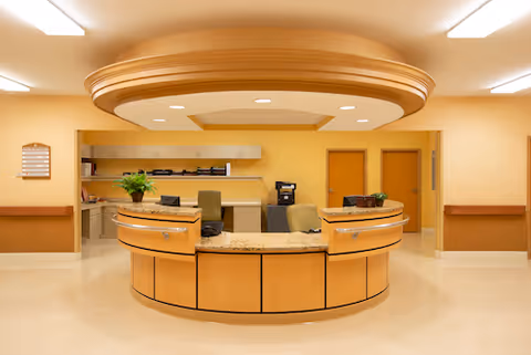 Reception desk area in a senior living facility with a curved wooden desk, granite countertop, office chairs, and cabinets in the background. The walls and ceiling are painted in warm beige tones with recessed lighting.