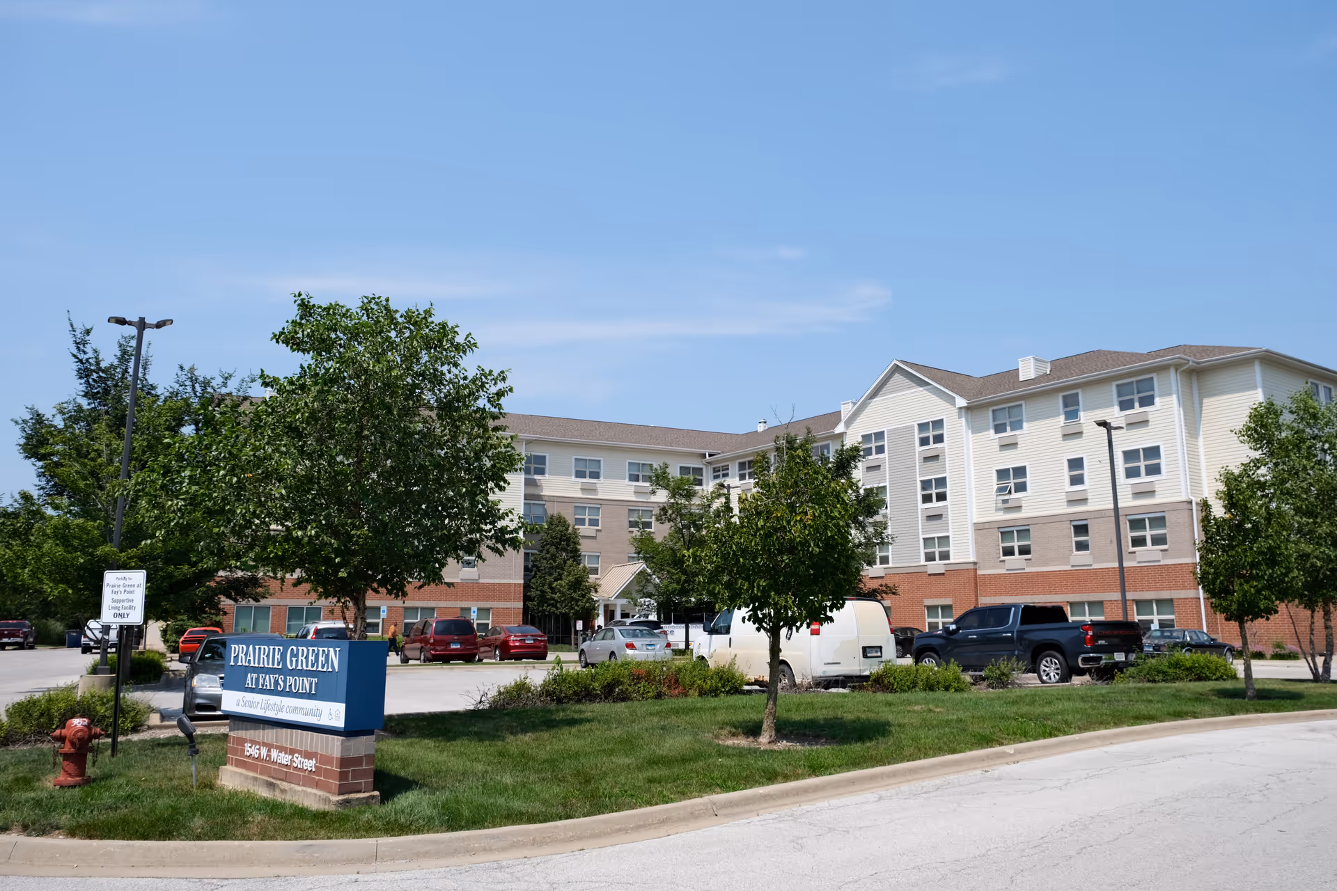Front view of a multi-story senior living building with a parking lot, trees, cars, and a sign reading 'Prairie Green at Fay's Point' under a clear blue sky.