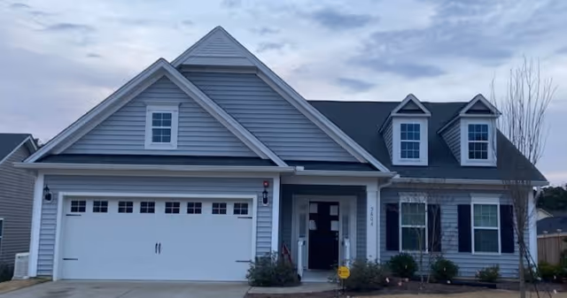 Exterior front view of a single-story residential building with gray siding, a two-car garage, and a small front porch. The house has multiple windows with black shutters and a driveway leading up to the garage. The sky is partly cloudy.