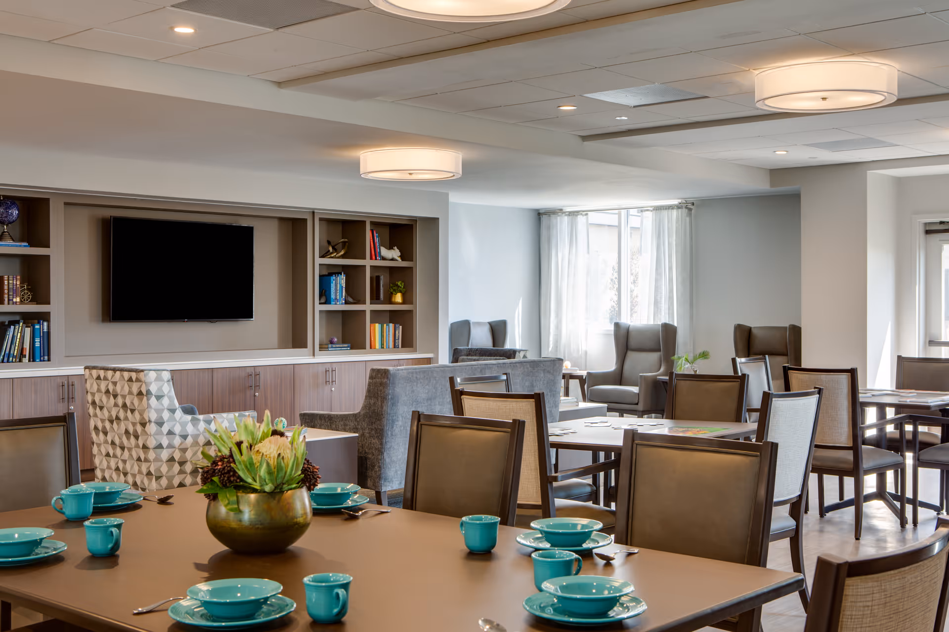 A bright and spacious common area in a senior living facility with multiple tables set with turquoise dishes and cups. Comfortable chairs and armchairs are arranged around the room, with a large flat-screen TV mounted on the wall above built-in shelves filled with books and decorative items. Large windows with sheer curtains allow natural light to fill the space.