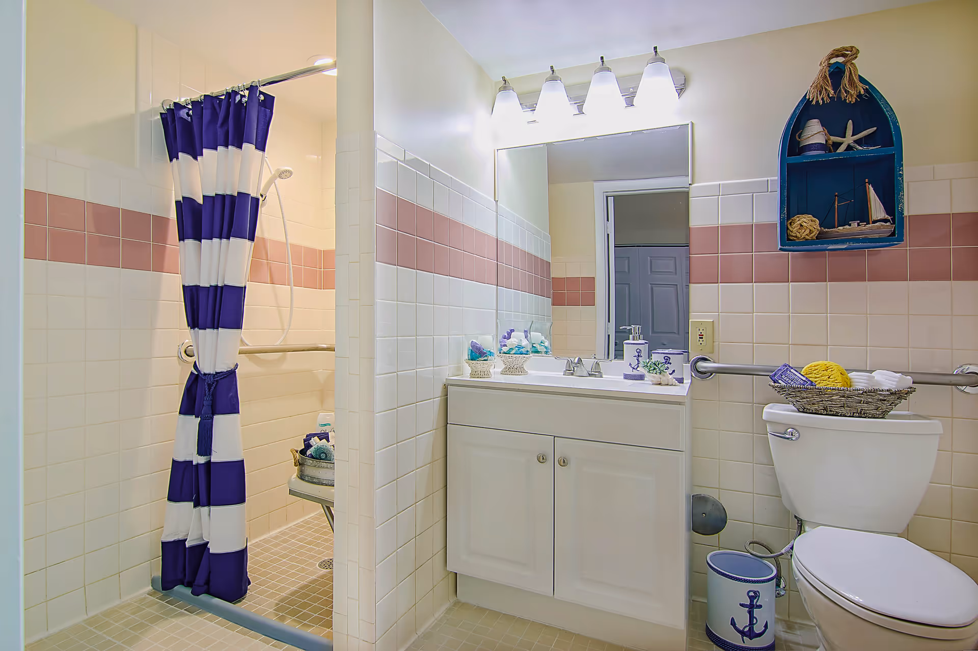 A bathroom with a white vanity and sink, a large mirror with four lights above it, a toilet with a basket of towels on top, and a walk-in shower with a blue and white striped curtain. The walls are tiled with white and pink tiles, and there is a blue nautical-themed wall shelf with decorative items above the toilet.