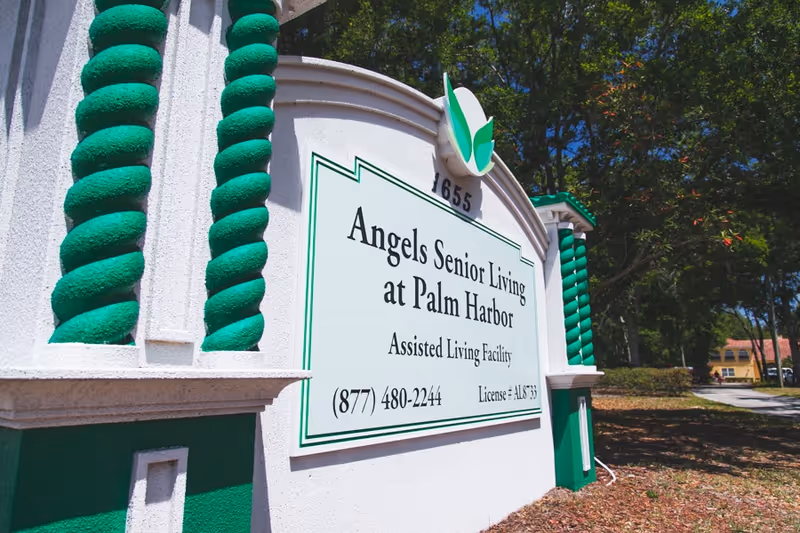 Outdoor sign for Angels Senior Living at Palm Harbor, an assisted living facility, with green and white decorative pillars and trees in the background.
