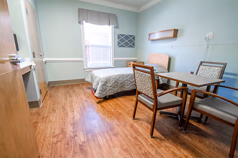 A nursing facility room with a single bed covered with a patterned bedspread, a window with blinds and a valance, a small table with four chairs, and light green walls with white trim.