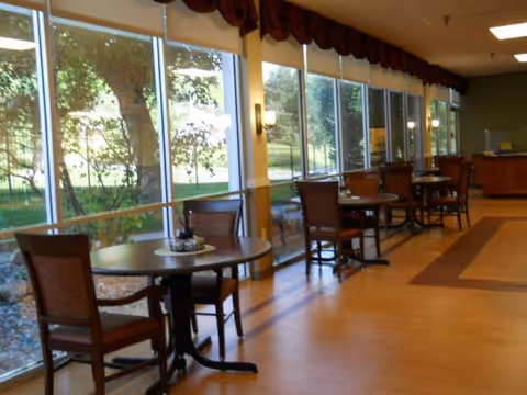 Interior of a senior living dining area with round tables by large windows.
