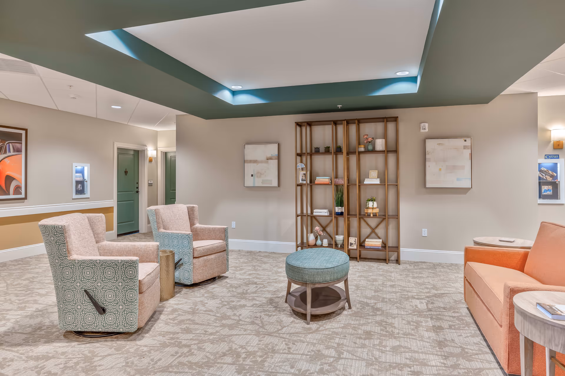 Communal lounge area with upholstered chairs, a round ottoman, shelving unit, and recessed ceiling lighting.