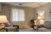 A cozy living room with two upholstered armchairs and a sofa, a wooden side table with a lamp, a potted plant, and a window with vertical blinds letting in natural light. The walls are decorated with simple molding and sconces.