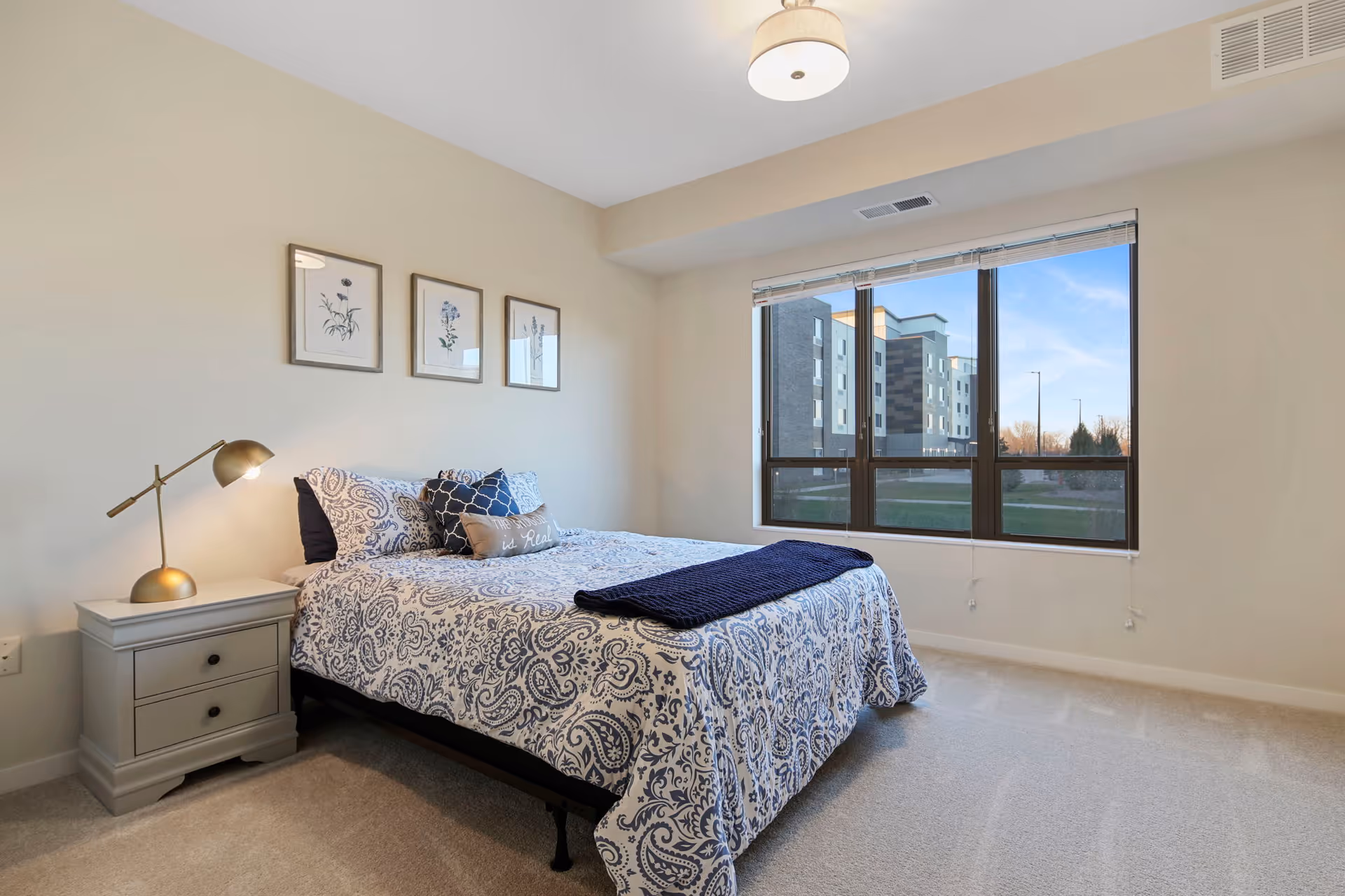 A neatly made bed with patterned blue and white bedding and decorative pillows in a bright bedroom. There is a nightstand with a gold lamp beside the bed, three framed botanical prints on the wall above the bed, and a large window showing an exterior view of a building and blue sky.