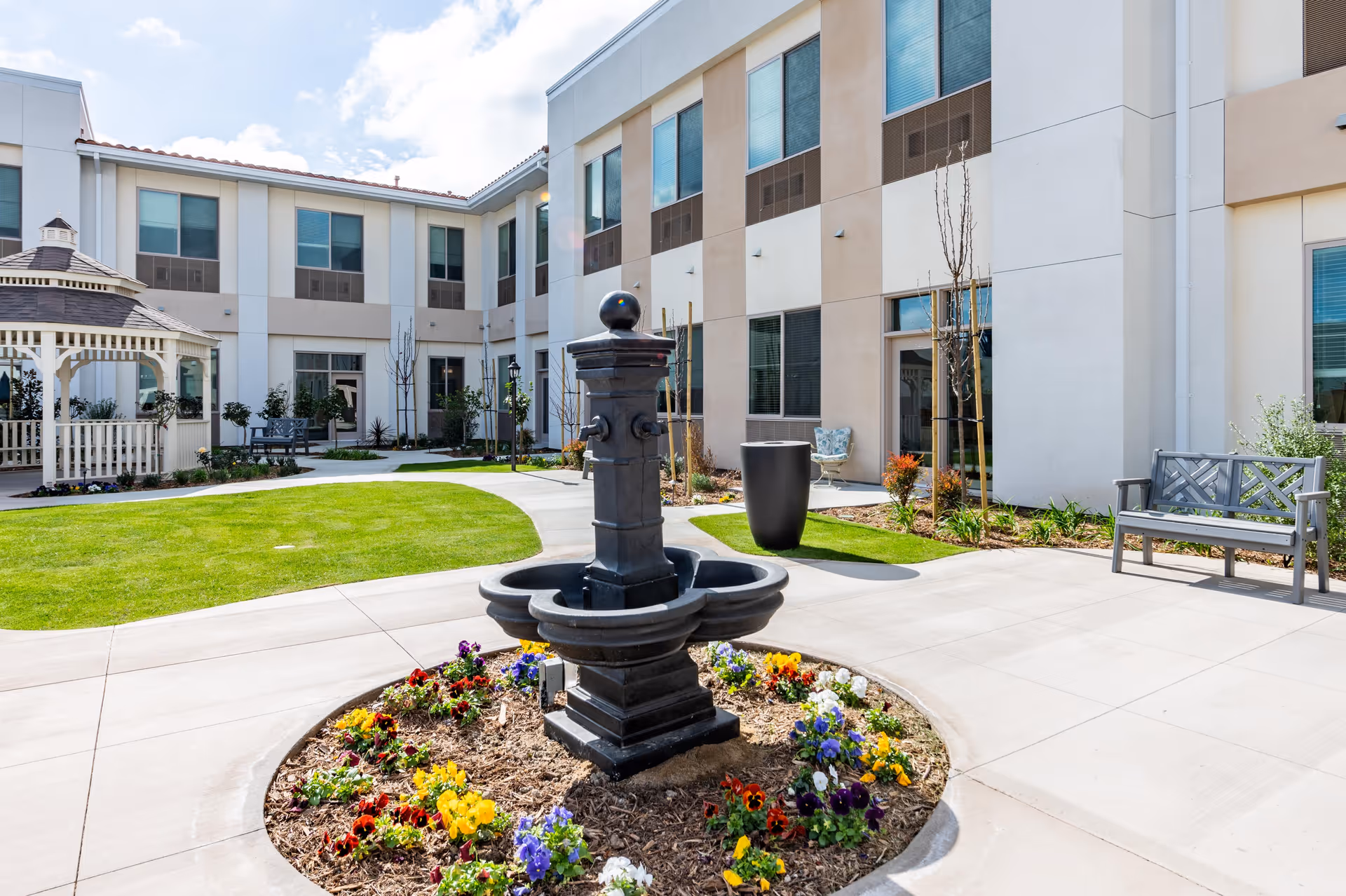 Outdoor courtyard area of a senior living facility with a central black fountain surrounded by colorful flowers, green grass, paved walkways, benches, and a white gazebo. The building has two stories with multiple windows and light-colored walls.