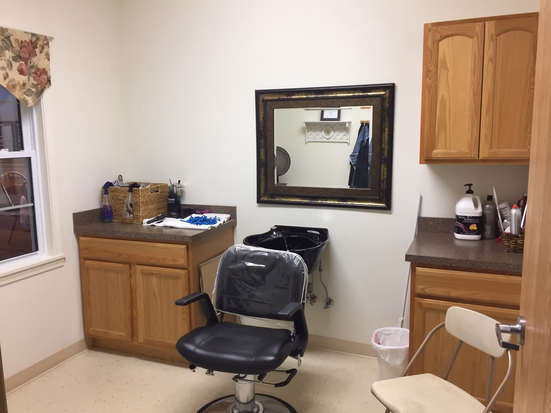 Interior of a small hair salon or grooming room with a black salon chair covered in plastic, a black wash basin mounted on the wall, a large framed mirror above the basin, wooden cabinets with various hair care products on the countertops, a window with floral valance, and a beige chair near the door.