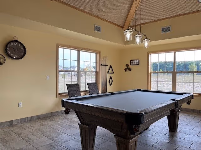 A room with a pool table in the center, two chairs positioned near two large windows, a wall clock, and a set of pool cues mounted on the wall. The room has a high ceiling with a hanging light fixture and tiled floor.
