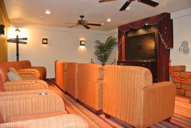 A cozy entertainment room with multiple orange upholstered armchairs arranged in rows facing a large flat-screen TV mounted on a wall with a decorative red curtain. The room has ceiling fans, wall sconces, a potted plant, and a striped carpet.