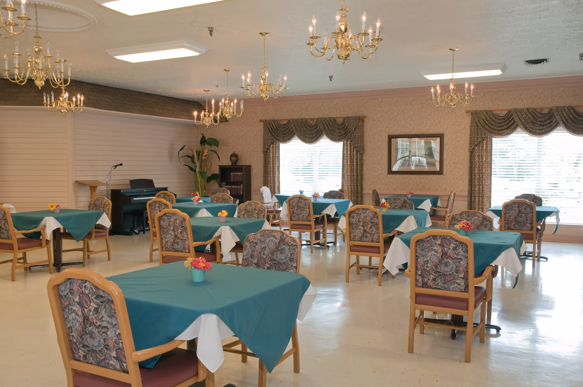 A dining room with multiple tables covered in teal tablecloths and white underlays, each table decorated with a small flower arrangement. The room has floral upholstered chairs, large windows with patterned curtains, chandeliers hanging from the ceiling, and a piano with a microphone stand in the corner.