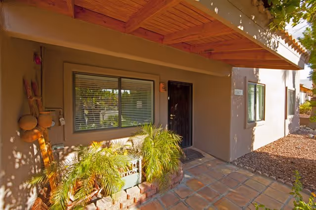 Exterior view of a residential building entrance with a wooden door, a window with blinds, a small garden bed with green plants, and a tiled walkway under a wooden awning.