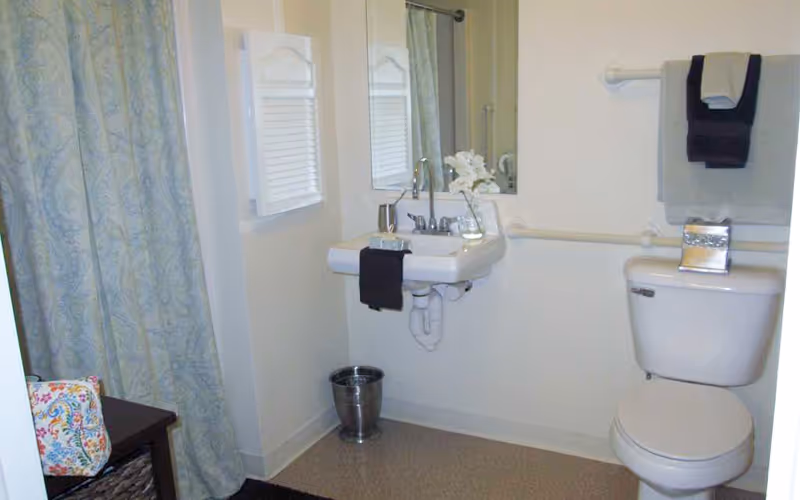 A clean bathroom with a white toilet and a wall-mounted sink. There is a mirror above the sink, a small vase with white flowers on the sink, and a black hand towel hanging from the sink. A towel rack above the toilet holds two folded towels, one black and one gray. A shower curtain with a light blue pattern is visible on the left side, and a small trash can is placed on the floor beneath the sink.