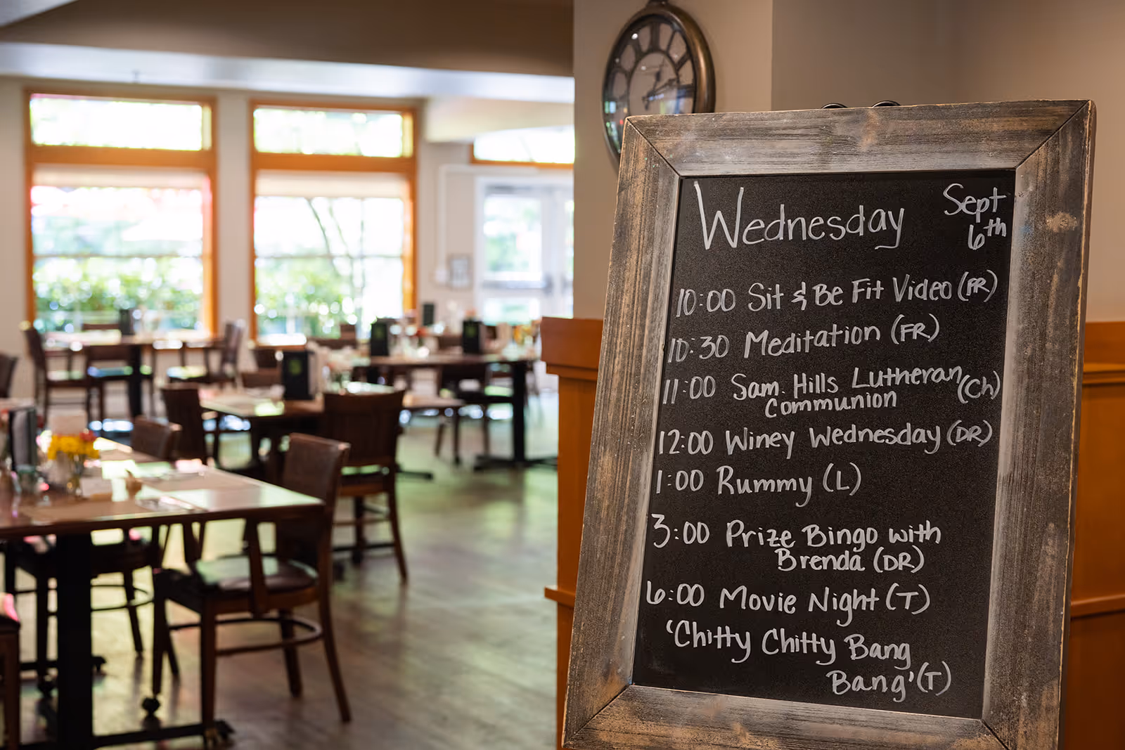 A dining room with several wooden tables and chairs, set with menus and small flower vases. In the foreground, there is a wooden-framed chalkboard displaying a schedule of activities for Wednesday, September 6th, including Sit & Be Fit Video, Meditation, Lutheran Communion, Winey Wednesday, Rummy, Prize Bingo, and Movie Night featuring 'Chitty Chitty Bang Bang'. Large windows in the background let in natural light and show greenery outside.