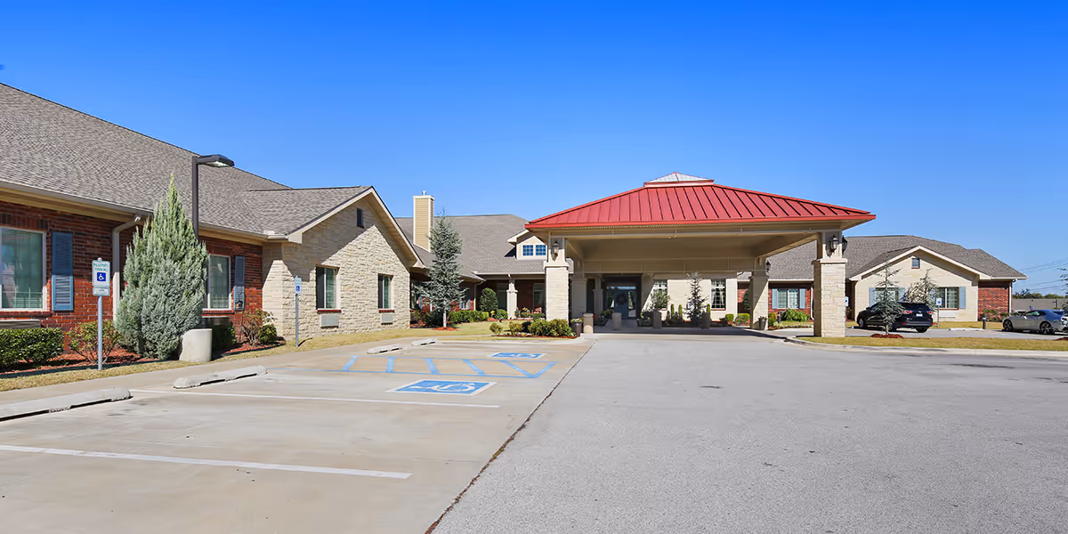 Exterior view of a senior living facility with a covered entrance featuring a red roof. The building has a combination of brick and stone walls, with several windows and landscaped greenery around. There are designated handicap parking spaces in front of the entrance under a clear blue sky.
