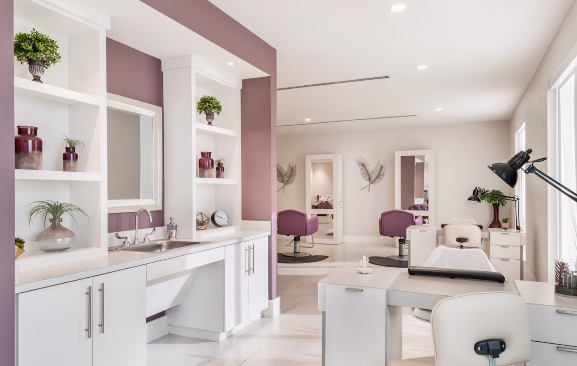 A bright and modern salon area with white cabinetry and countertops, a sink, decorative plants and vases on shelves, purple accent walls, salon chairs in front of mirrors, and a manicure table with lamps near large windows.