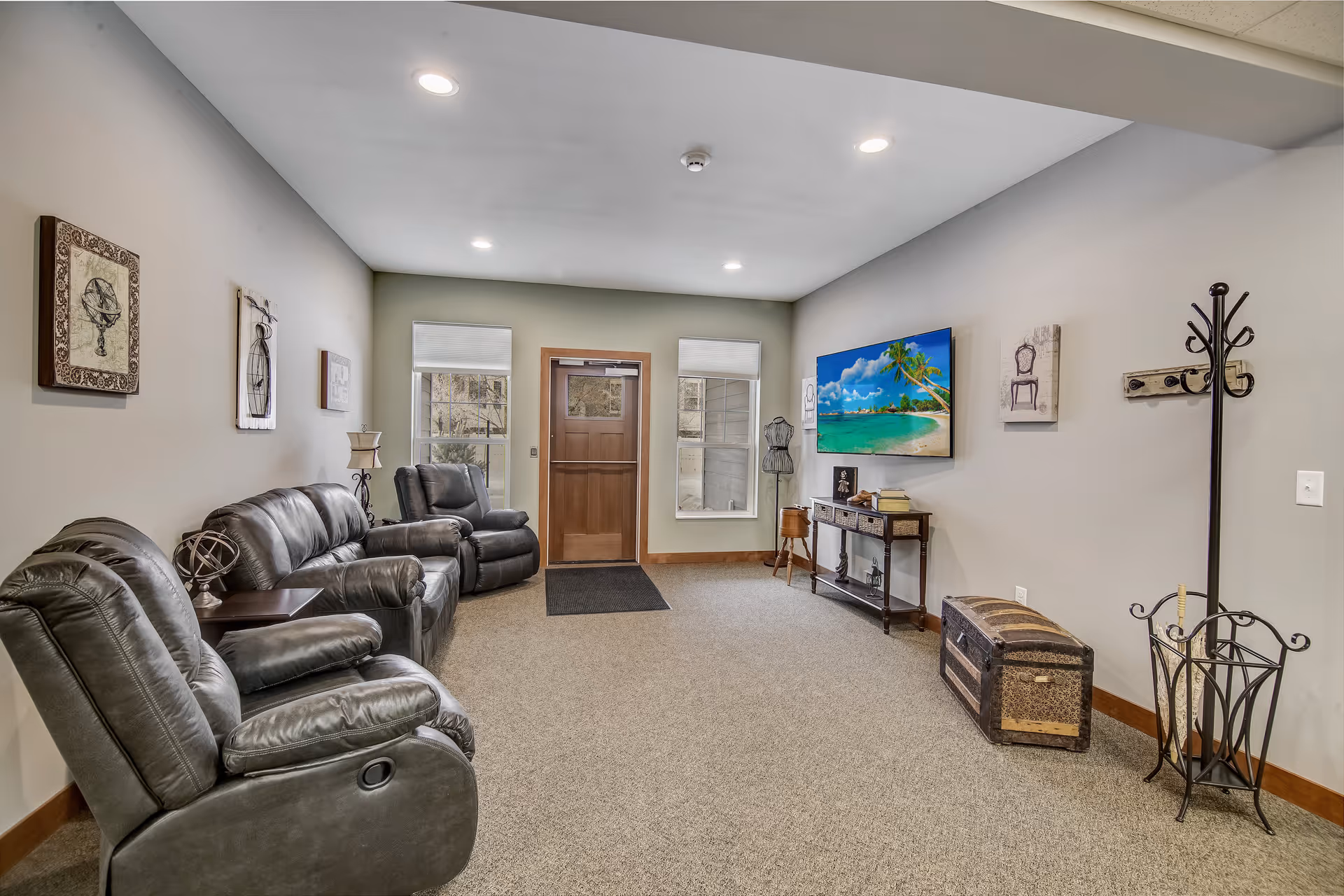 A senior living facility common area with three black leather recliners arranged along the left wall. The room has beige walls with framed artwork, a wooden door with two windows on either side, and a flat-screen TV mounted on the right wall displaying a tropical beach scene. Below the TV is a decorative console table with books and ornaments. There is a coat rack and an umbrella stand near the right wall, and a carpeted floor throughout the room.