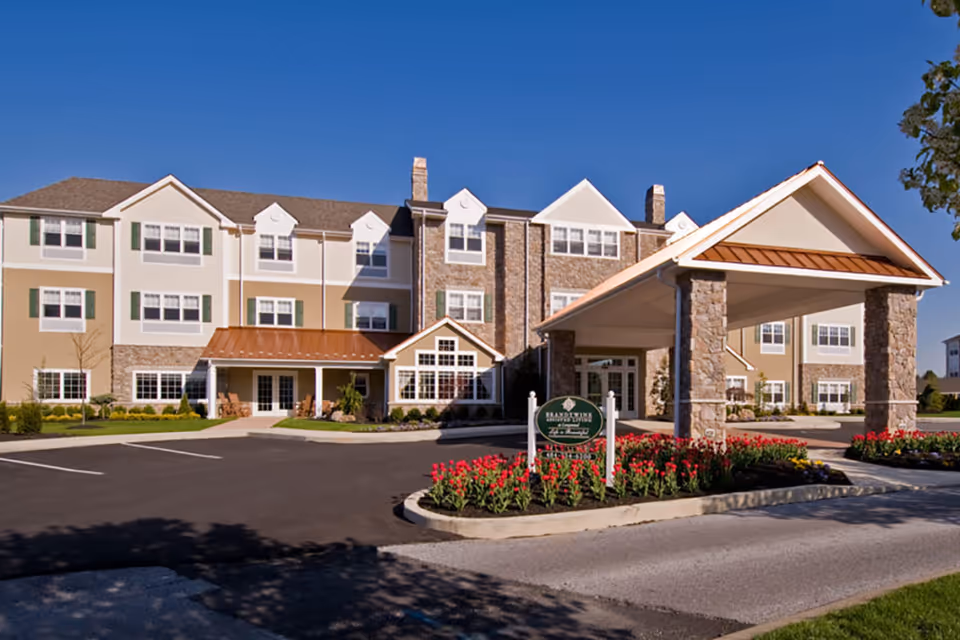 Exterior view of a three-story senior living facility building with beige and stone facade, multiple windows, and a covered entrance with stone pillars. There is a landscaped area with red flowers and a sign that reads 'Brandywine Longwood by Monarch'. The sky is clear and blue.