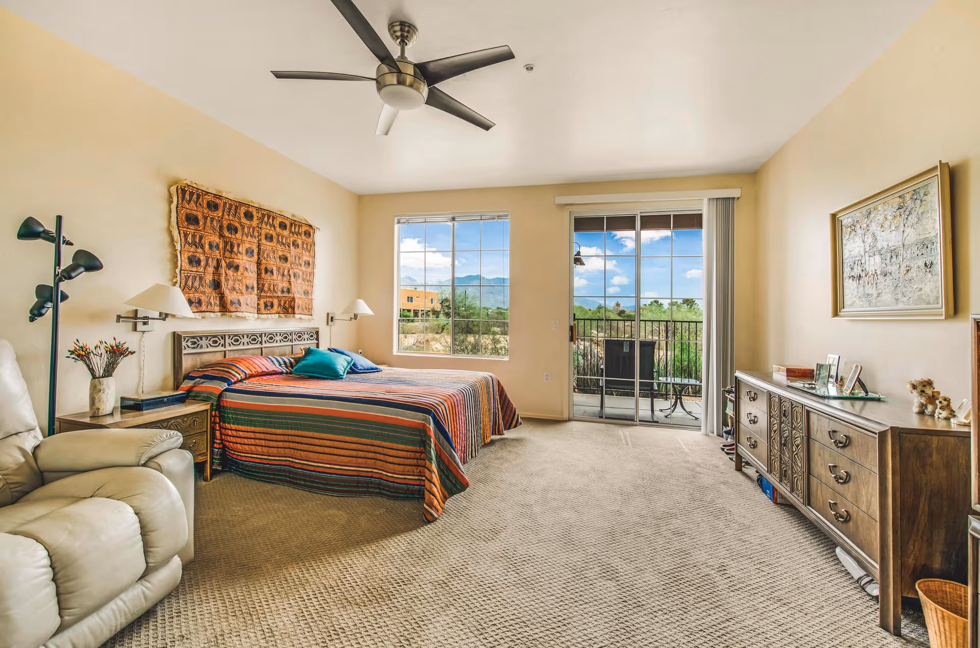 A spacious bedroom with a large bed covered in a colorful striped bedspread, two pillows, and a decorative wall hanging above the headboard. There is a beige recliner chair to the left, a wooden nightstand with a vase of flowers, and a floor lamp. The room has a large window and a sliding glass door leading to a balcony with outdoor furniture. A wooden dresser with framed pictures and decorative items is on the right wall, and a ceiling fan is mounted above.