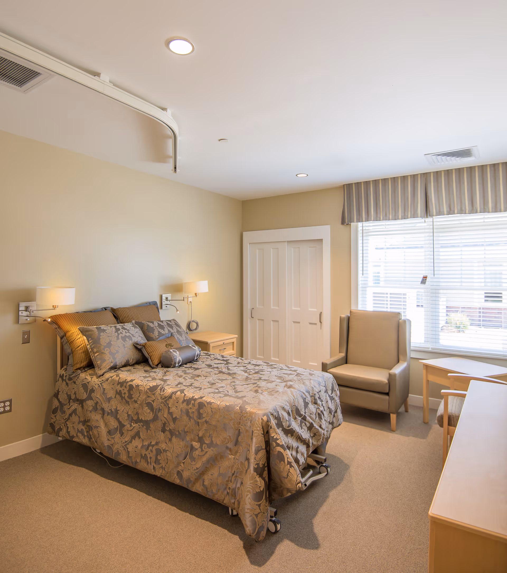 A neatly made bed with patterned bedding in a well-lit bedroom. The room features beige walls, a window with blinds and a striped valance, a comfortable armchair, a wooden nightstand with a lamp, and a closet with white sliding doors.