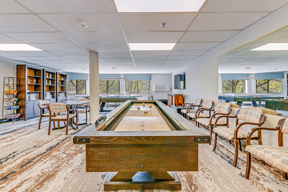 A bright and spacious recreational room in a senior living facility featuring a shuffleboard table in the center, several cushioned chairs with wooden armrests arranged along the walls, a table with chairs in the background, large windows letting in natural light, and shelves with decorative items and games.