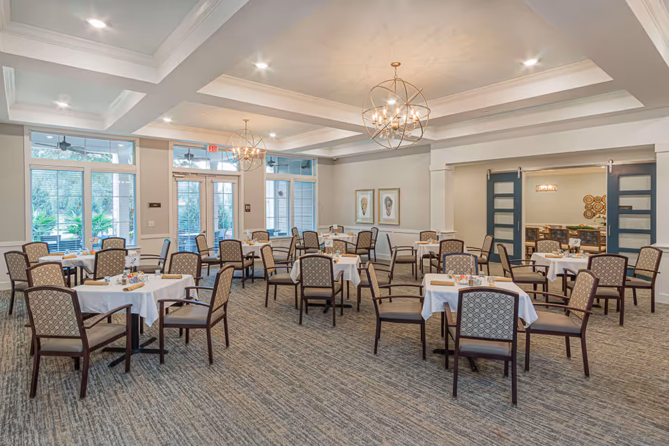 Spacious dining room with several tables draped in white linens and patterned chairs beneath modern chandeliers.