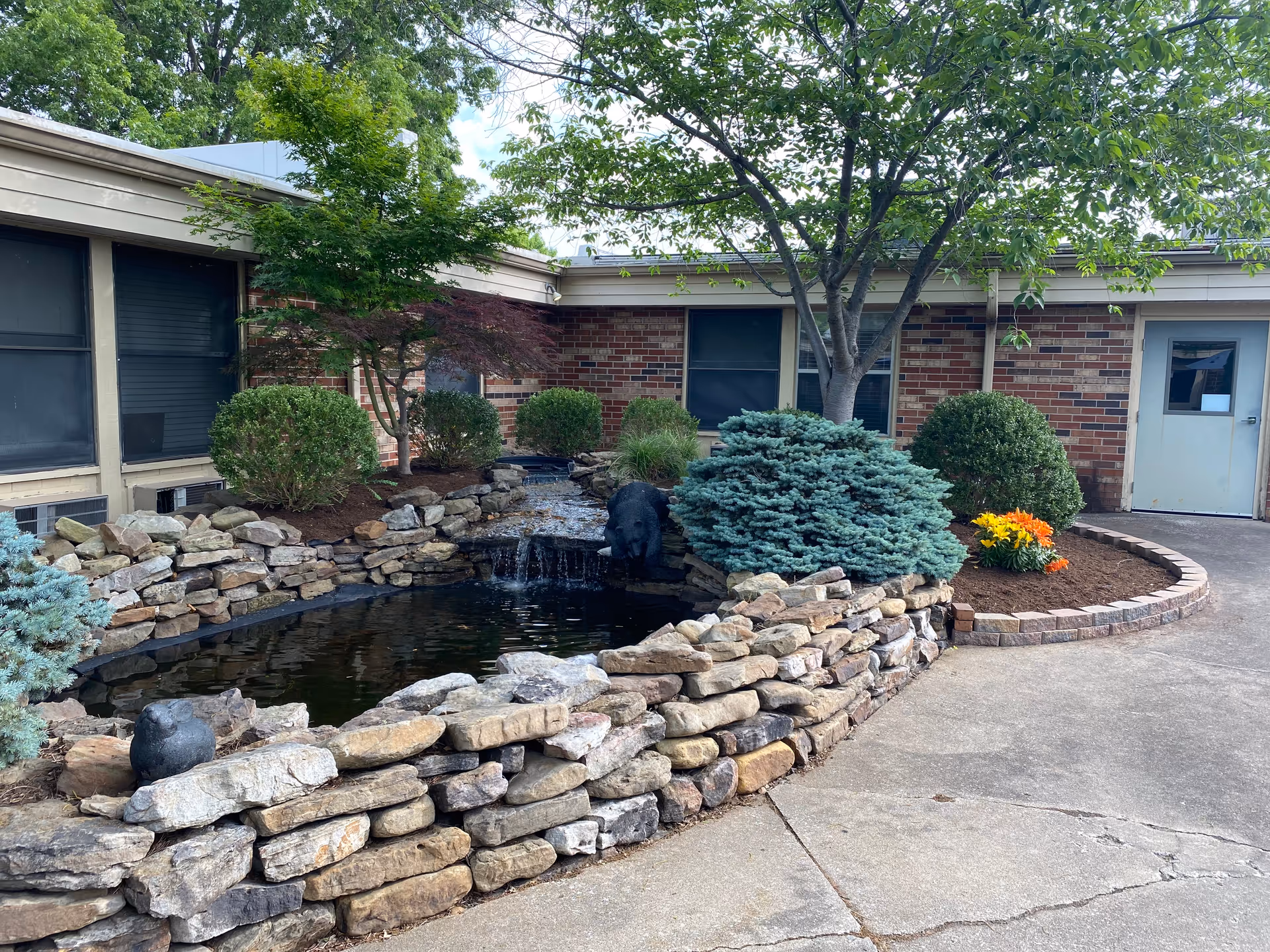 Outdoor courtyard area with a small pond surrounded by a stone wall, various shrubs, a tree, and a brick building with windows and a door in the background.