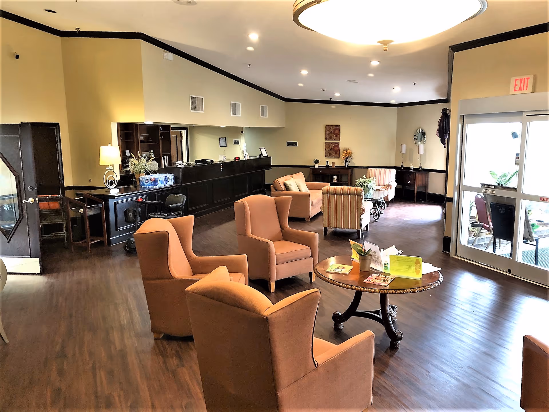 A spacious senior community lounge area with several comfortable armchairs arranged around a round wooden table. The room features wood flooring, a reception desk with a lamp and decorative plants, and a glass door leading outside. The walls are painted light yellow with dark trim, and the ceiling has recessed lighting and a large circular light fixture.