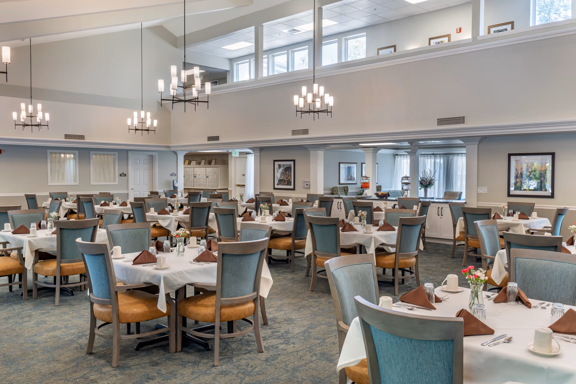 A spacious dining room with multiple round tables covered with white tablecloths, each set with brown folded napkins, cups, and silverware. The room has high ceilings with modern chandeliers, large windows near the ceiling allowing natural light, and light-colored walls adorned with framed artwork. Chairs with blue upholstery and wooden frames surround each table.