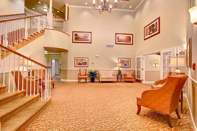 Spacious senior living facility common area with patterned carpet, beige walls, and high ceilings. The room features a staircase with white railings on the left, several upholstered chairs and sofas arranged along the walls, framed artwork, a chandelier, and table lamps providing warm lighting.