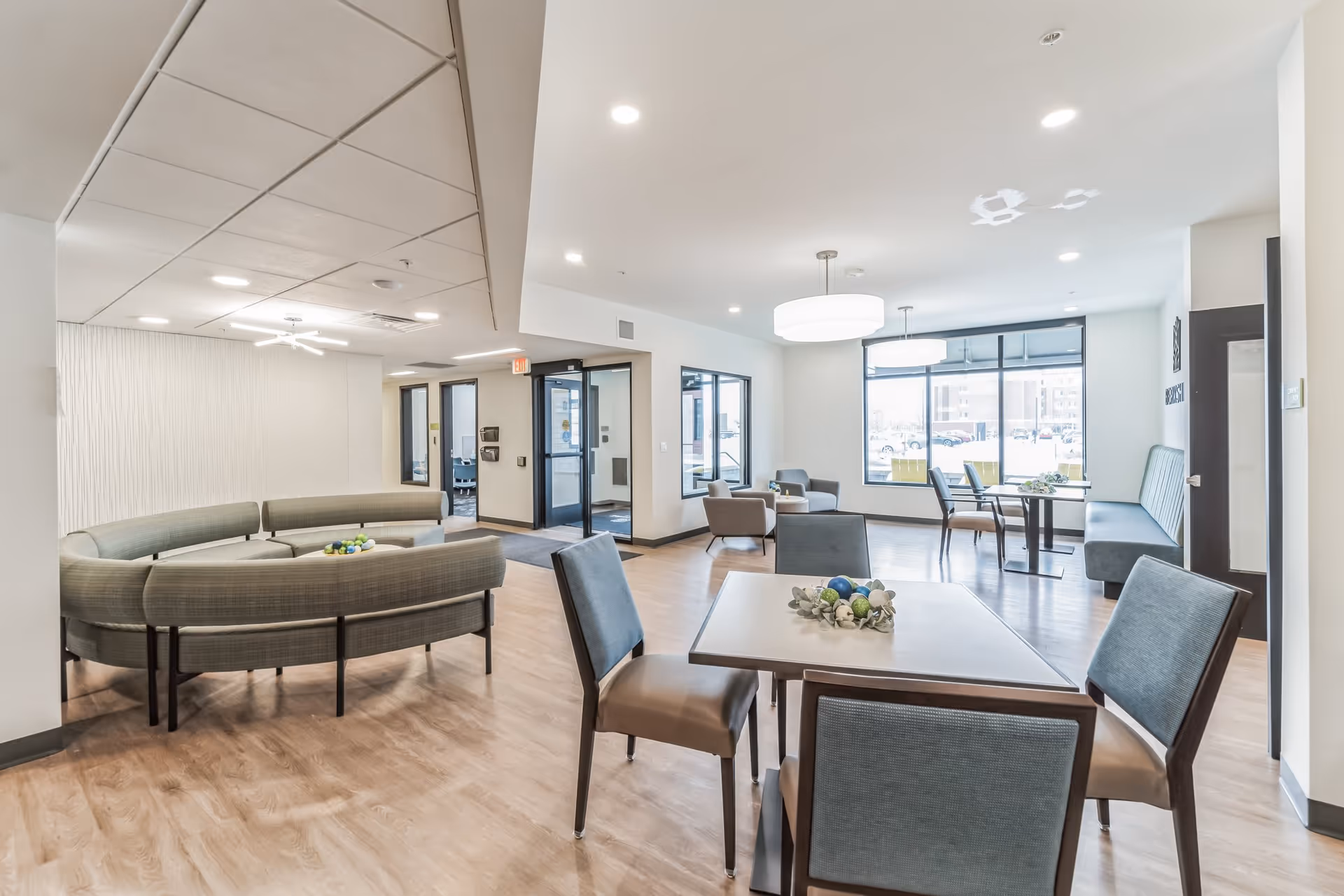 A bright and modern common area in Berkshire Ashwaubenon Apartments featuring a circular green cushioned bench, several tables with chairs, and large windows letting in natural light. The space has light wood flooring, white walls, and ceiling lights, creating an inviting atmosphere.