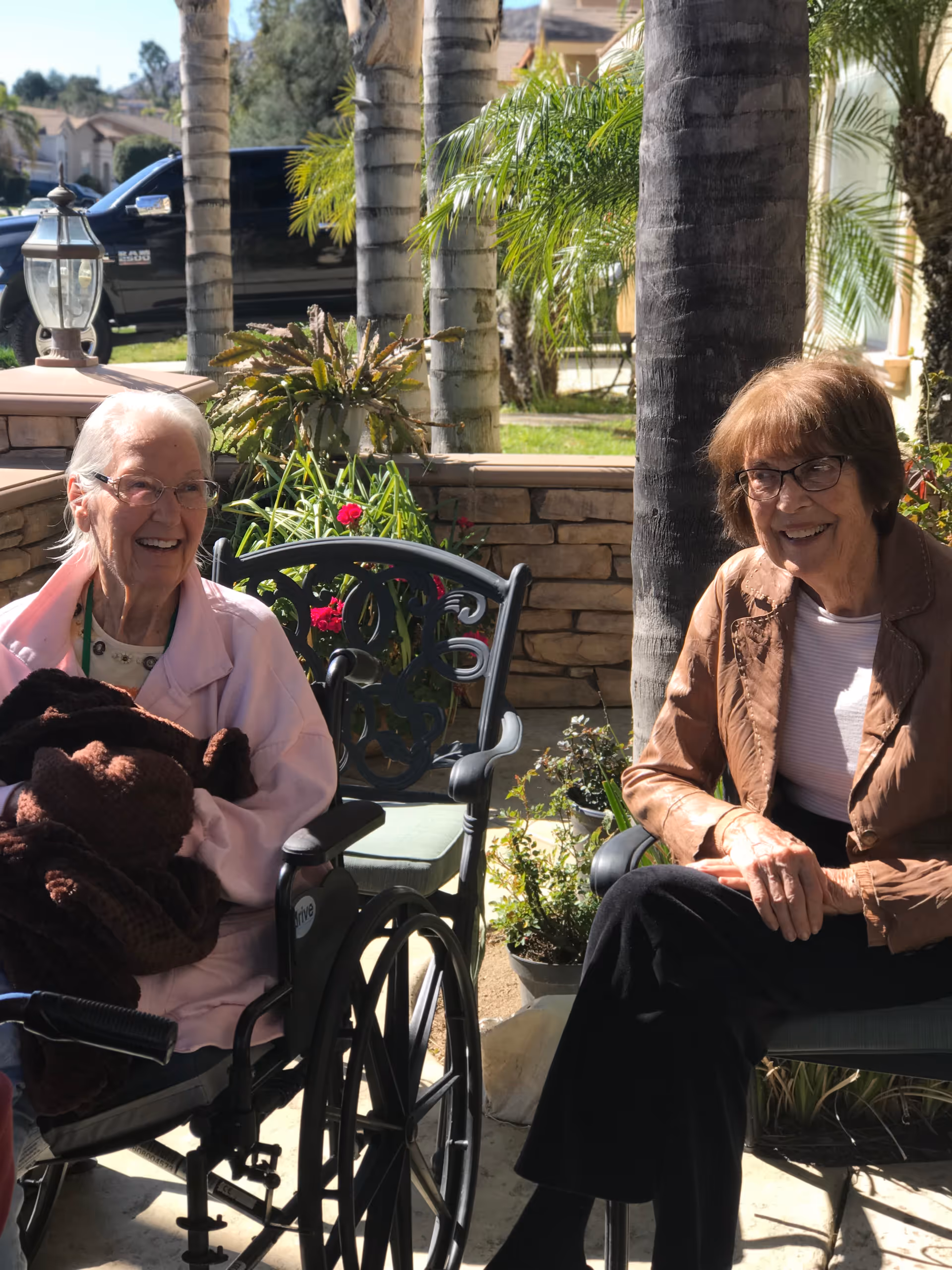 Two elderly women sitting outdoors on a sunny day. One woman is in a wheelchair wearing a light pink jacket and holding a brown blanket, smiling. The other woman is seated on a chair wearing glasses, a brown jacket, and black pants, also smiling. They are surrounded by palm trees, plants, and a stone wall with a residential neighborhood visible in the background.