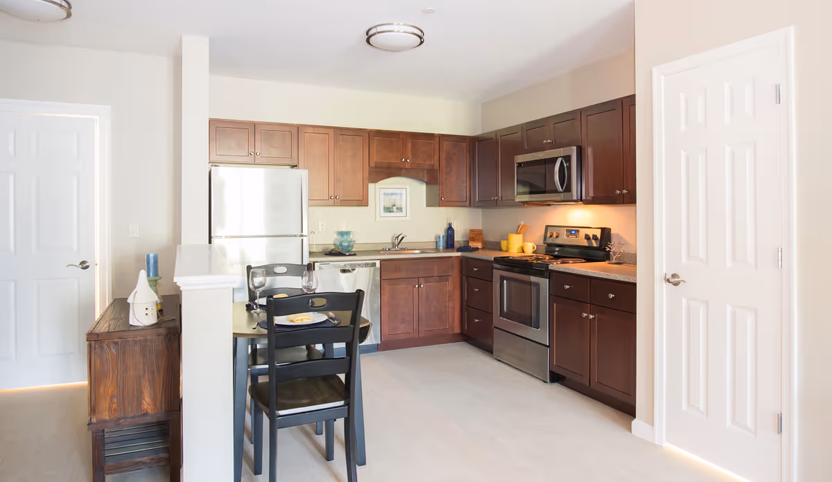 A modern kitchen with dark wood cabinets, stainless steel appliances including a refrigerator, dishwasher, stove, and microwave. There is a small dining table with two black chairs set with plates and glasses. The room has light-colored walls and flooring, with two white doors visible on either side.
