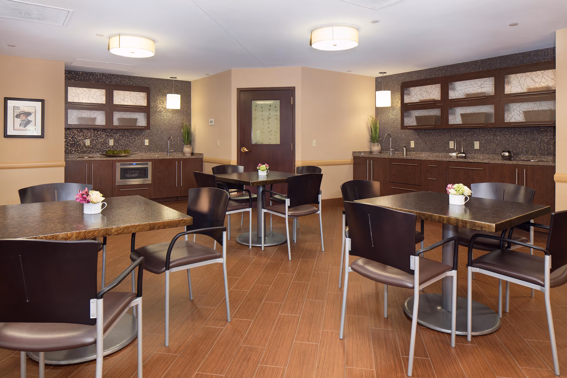 A dining area with several square tables and dark chairs arranged neatly. Each table has a small flower arrangement in a white mug. The room features wood flooring, beige walls, and a kitchenette area with dark wood cabinets, a microwave, and a sink. Two ceiling lights and two hanging pendant lights illuminate the space.