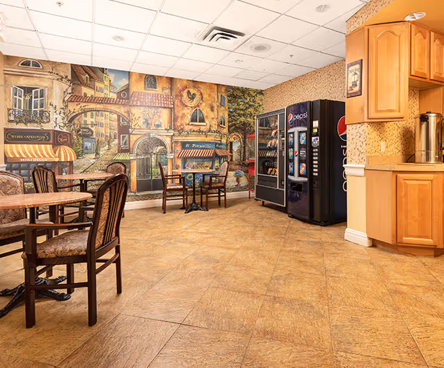 A dining area with several tables and chairs, a vending machine with Pepsi branding, and a mural on the wall depicting a European street scene with buildings and storefronts. The floor is tiled, and there are wooden cabinets on the right side.
