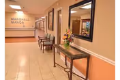 A clean and well-lit hallway in Marshall Manor Nursing & Rehab Center with tiled floors, beige and peach colored walls, a row of chairs along one side, a decorative table with a flower arrangement and a large mirror above it, and a sign on the far wall that reads 'Marshall Manor'.