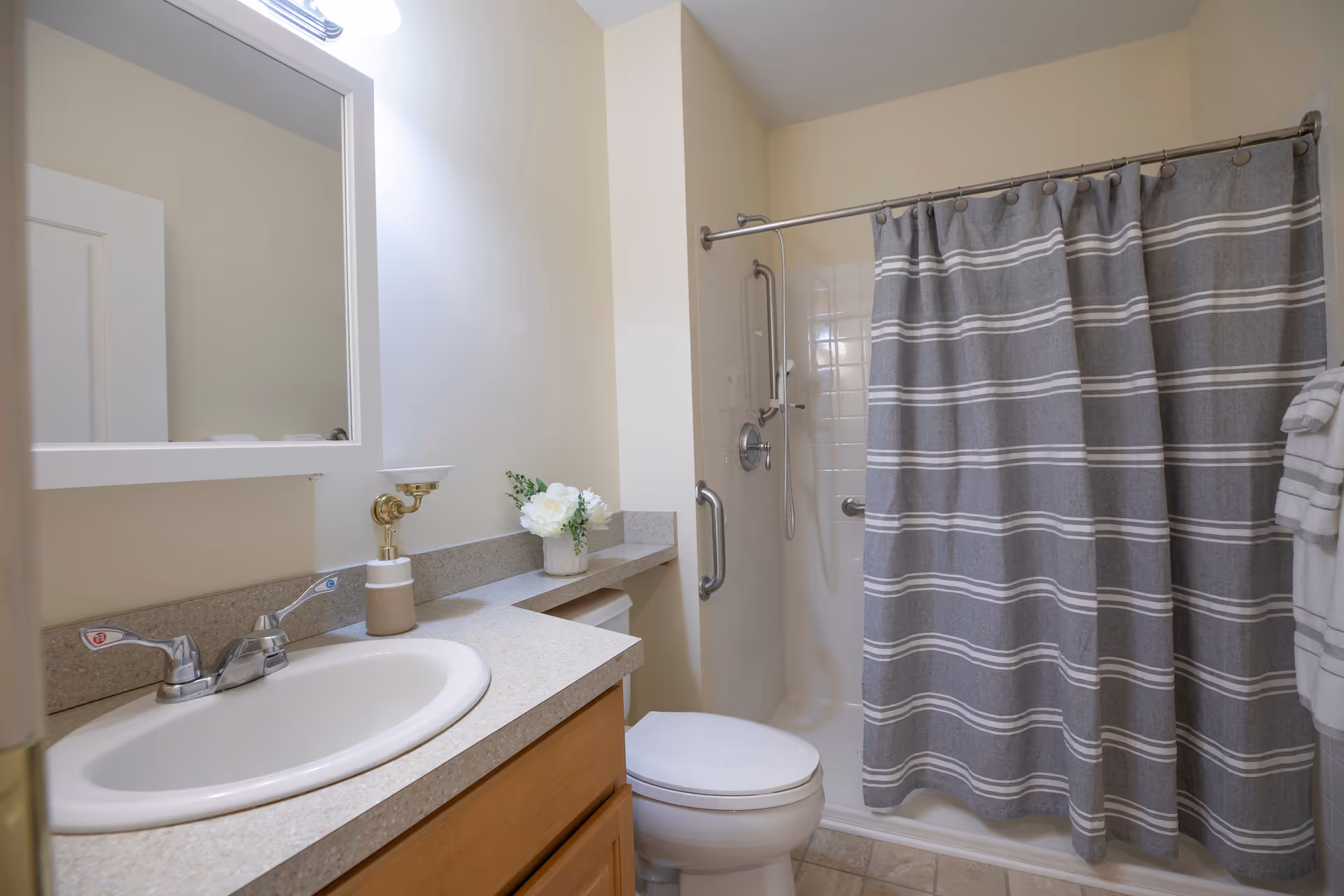 A clean bathroom featuring a white sink with silver faucet handles, a countertop with a soap dispenser and a small vase with white flowers, a toilet, and a shower area with a gray and white striped curtain. The walls are light-colored, and there is a mirror above the sink with a light fixture above it.