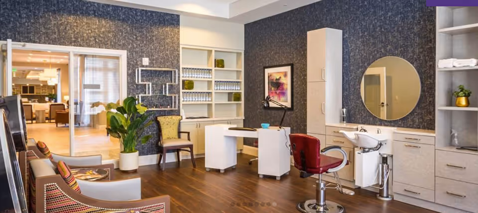 Interior view of a stylish salon area in a senior living facility with a red salon chair in front of a round mirror and sink, a white desk, a chair, built-in shelves with decorative items, and a seating area with armchairs and cushions. The room has wooden flooring and dark textured wallpaper.