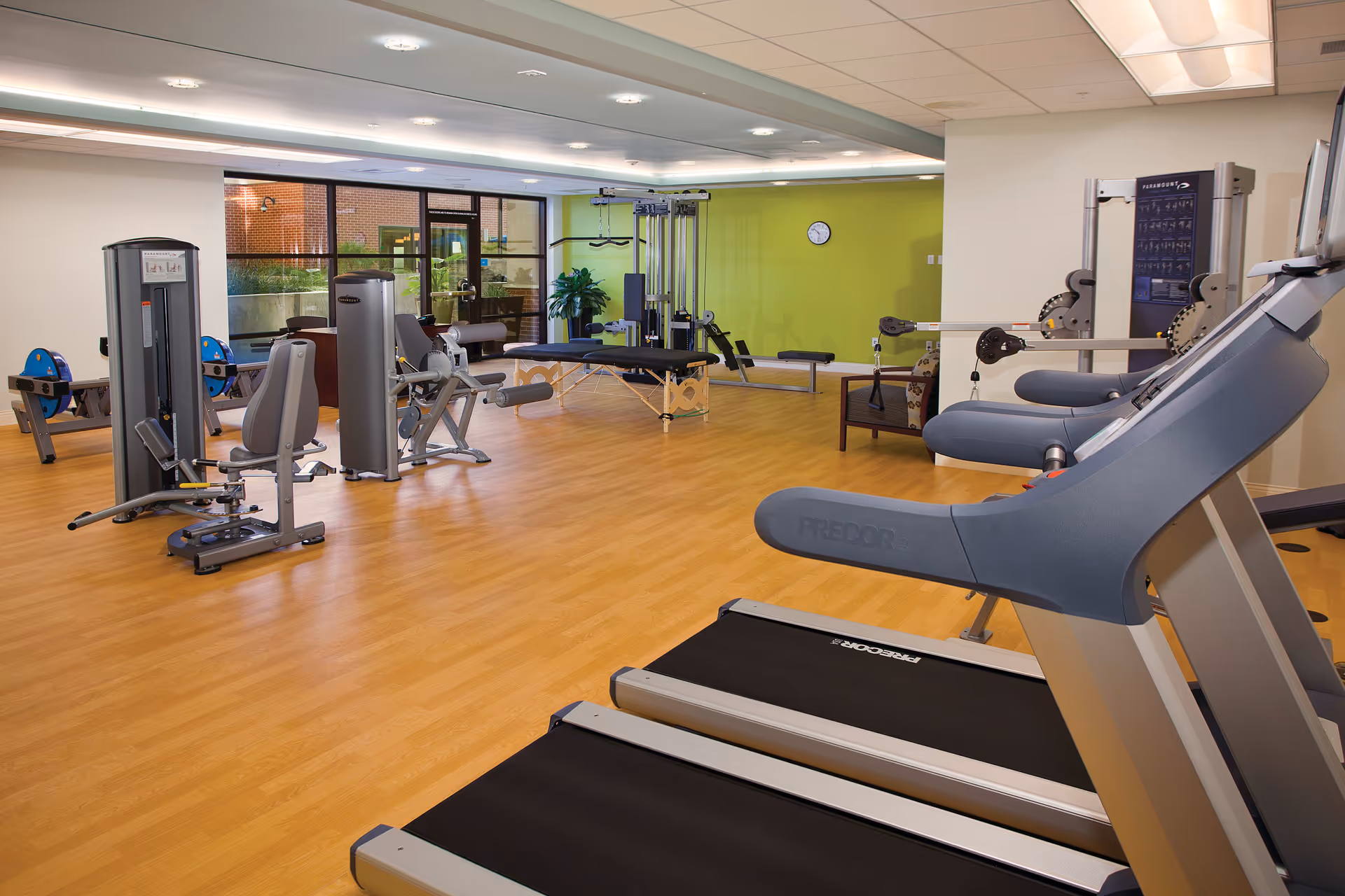 A spacious fitness room with various exercise equipment including treadmills, weight machines, and benches. The room has wood flooring, a green accent wall with a clock, large windows letting in natural light, and a seating area with a chair.
