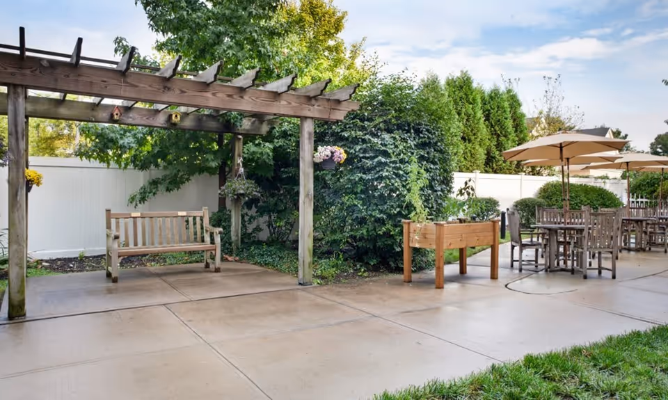Outdoor patio area with a wooden pergola and bench on the left, surrounded by greenery and hanging flower pots. To the right, there are several wooden tables and chairs with large beige umbrellas providing shade. The area is paved and enclosed by a white fence with trees and bushes in the background.
