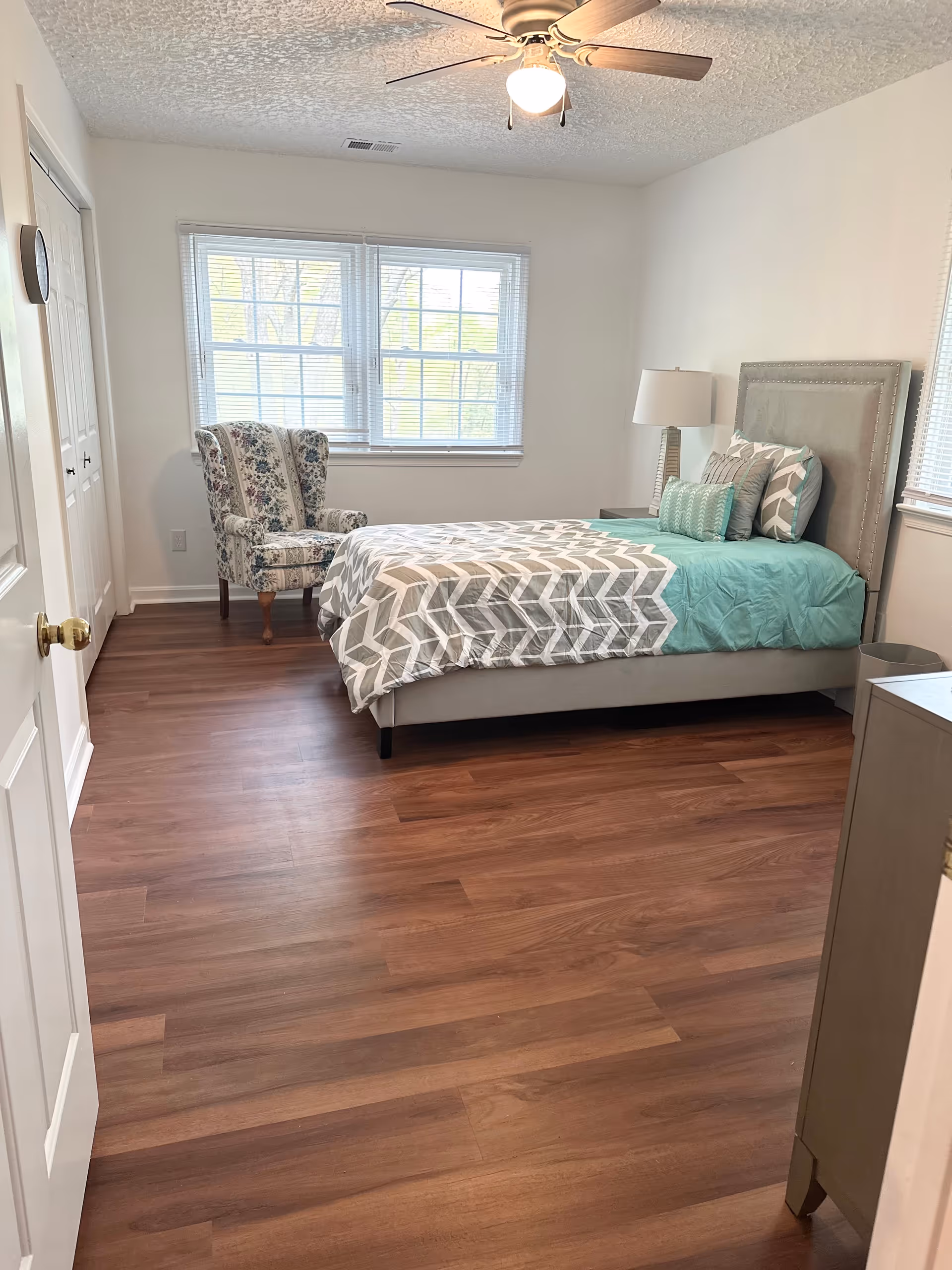 Bright furnished bedroom with a single bed, patterned armchair by a window, ceiling fan, and wood-look flooring.