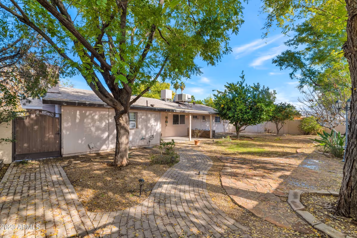 Backyard of a single-story house with a curved paved walkway, mature trees, and a fenced yard.