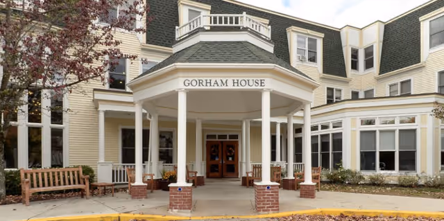 Front exterior view of Gorham House, a multi-story building with beige siding and white trim. The entrance features a covered porch supported by white columns and a sign above reading 'GORHAM HOUSE'. There are benches and some landscaping around the entrance area.