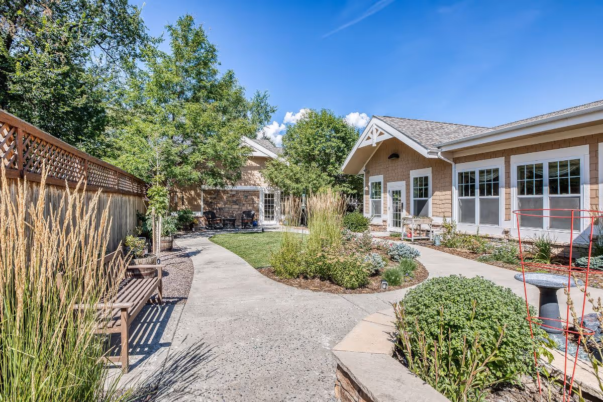 Sunny landscaped courtyard with a winding concrete path, benches, garden beds, and a single-story assisted living building.