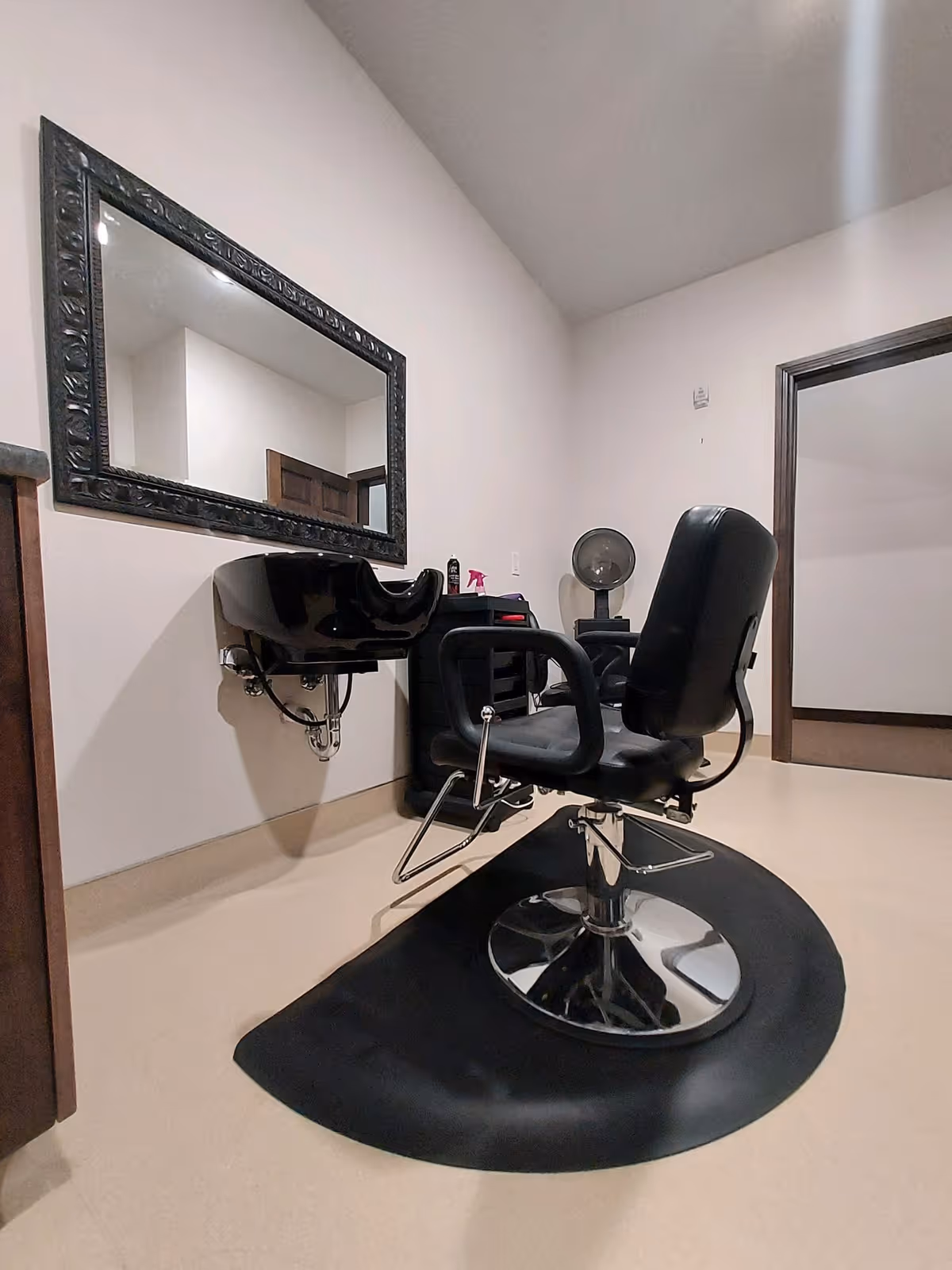 Interior view of a hair salon area with a black salon chair on a black mat, a black sink attached to the wall, a large decorative mirror, and a hair dryer in the background.