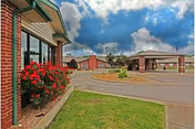 Front exterior of a brick building with a covered entrance, circular driveway, and flower beds beneath a cloudy sky.