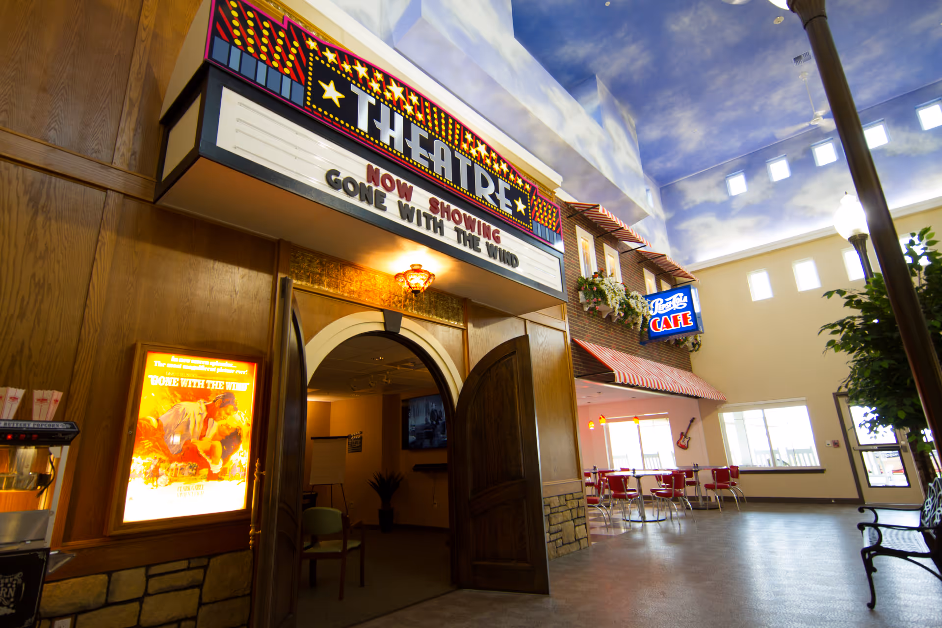 Interior of a senior living facility featuring a theater entrance with a marquee sign reading 'THEATRE NOW SHOWING GONE WITH THE WIND'. To the right, there is a cafe area with red chairs and tables under a sign that says 'Pete's Cafe'. The ceiling is painted with a sky and clouds motif, and the space is well-lit with natural light from high windows.